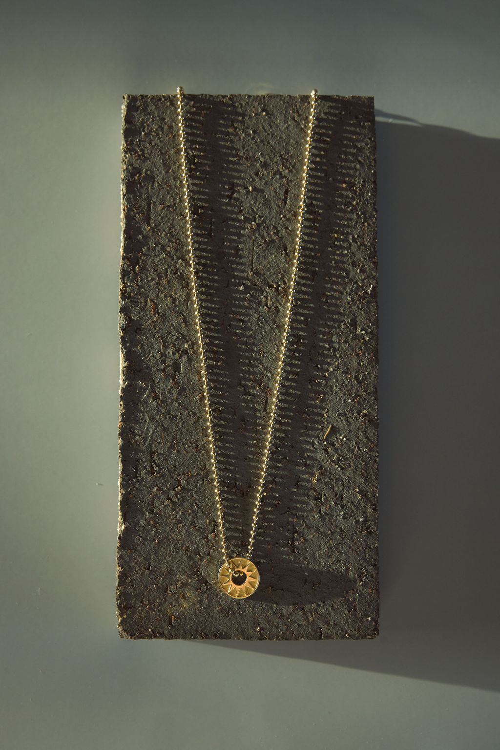 Gold ball chain necklace with round sun pendant displayed on a textured stone block